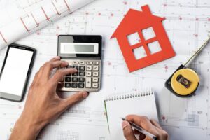 Hands using a calculator next to a red house cutout, blueprints, and a tape measure while estimating a home's value