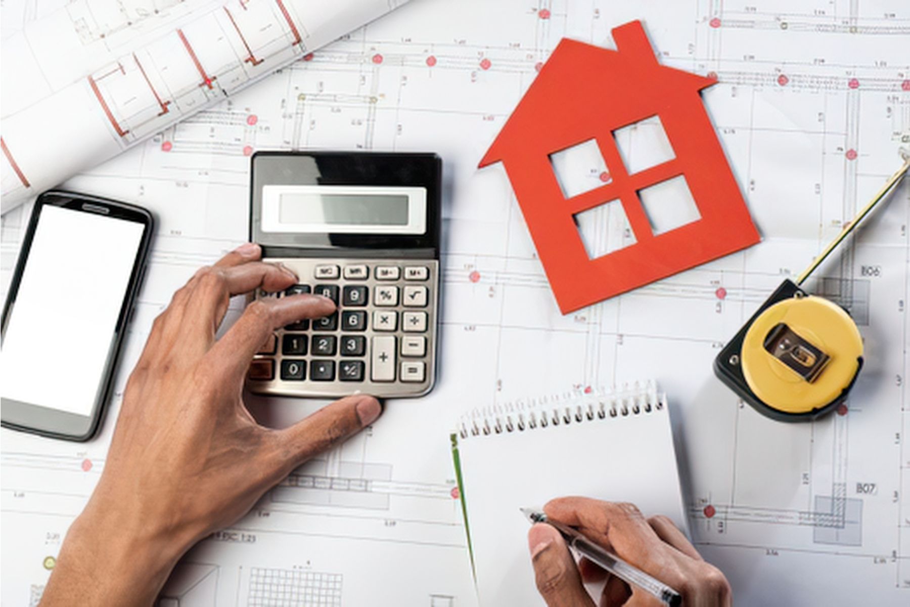 Hands using a calculator next to a red house cutout, blueprints, and a tape measure while estimating a home's value