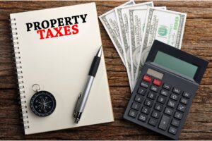 Notebook labeled Property Taxes next to cash, a calculator, and a compass, representing property tax proration in Indiana home sales.