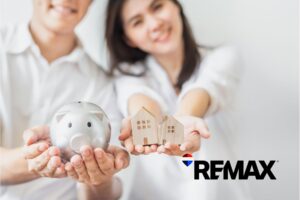 Young couple holding a piggy bank and two small wooden houses, saving for their first home in Plainfield, Indiana